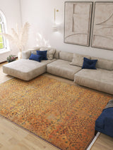 A modern living room features a large beige sectional, blue pillows, the Blossom Breeze - Washable Rug - JR1752 in orange, abstract wall art, pampas grass in a vase, daylight from a window, and light wood flooring.