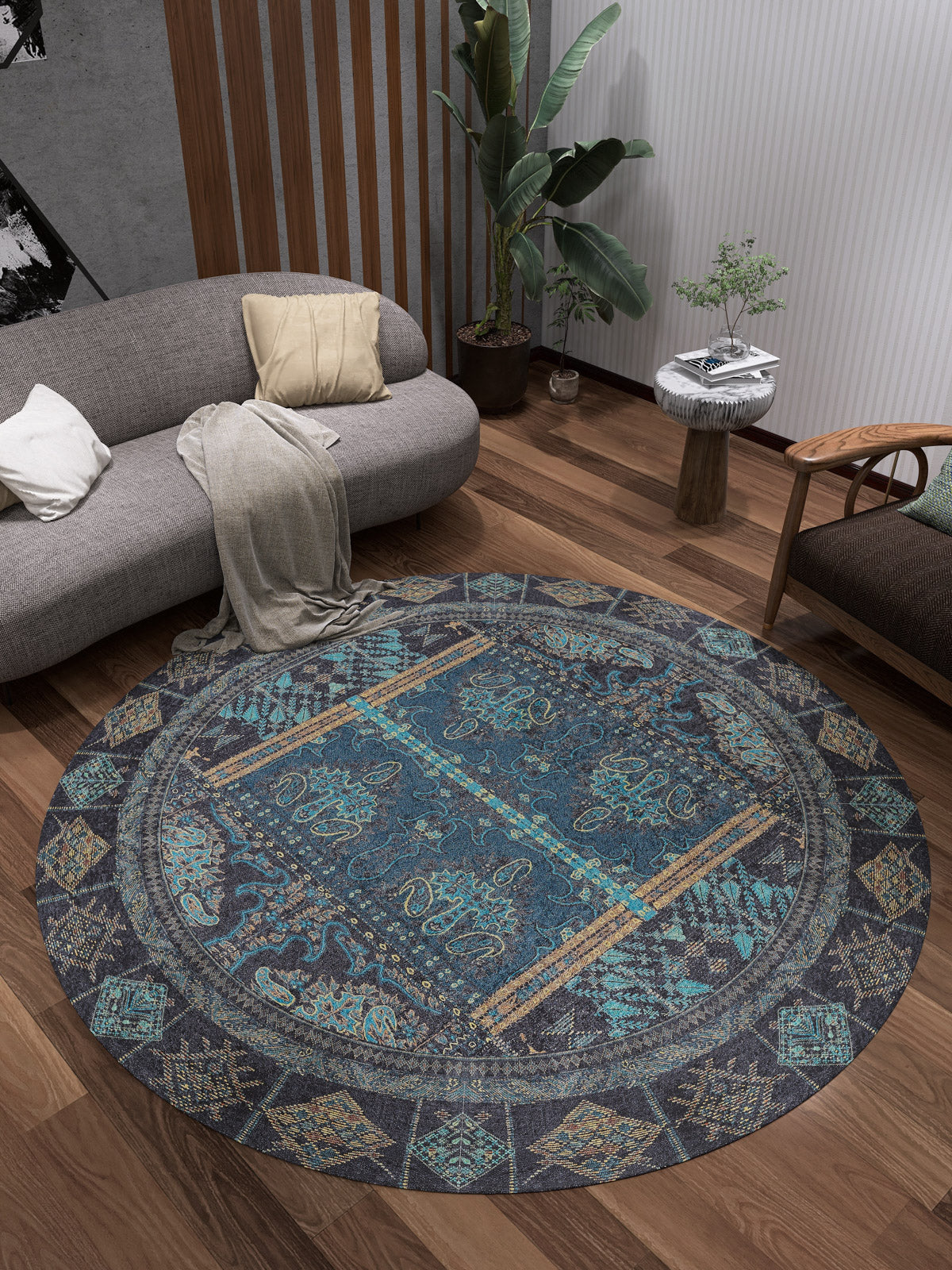 A modern living room with the Blossoming Bazaar - Washable Rug - JR1862, featuring a round blue pattern on wooden floors, a gray sofa with pillows and throw, mid-century armchair, plants, and minimalist decor—perfect for high-traffic areas.
