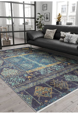 A modern living room features a dark sofa, decorative pillows, green plants, and large windows. The Blossoming Bazaar - Washable Rug - JR1862 (Custom Size) in blue with yellow and teal patterns covers the light wood floor—ideal for high-traffic areas.