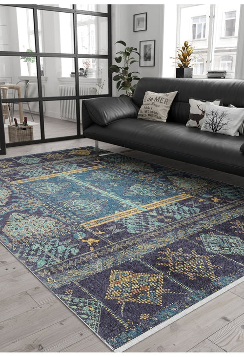 Load image 1 in gallery view. A modern living room features a dark sofa, decorative pillows, green plants, and large windows. The Blossoming Bazaar - Washable Rug - JR1862 (Custom Size) in blue with yellow and teal patterns covers the light wood floor—ideal for high-traffic areas.