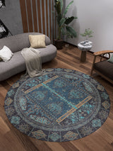 A modern living room with wooden floors features the Blossoming Bazaar - Washable Rug - JR1862 (Custom Size), ideal for high-traffic areas, along with a grey sofa, wooden armchair, large plant, and a side table topped with greenery.