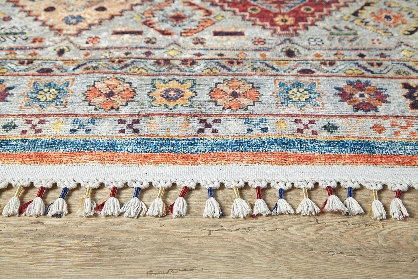 A close-up of the Bohemian Bouquet - Washable Rug - JR5090 (Custom Size), featuring intricate geometric and floral patterns with white fringe tassels, displayed on a light wooden floor.