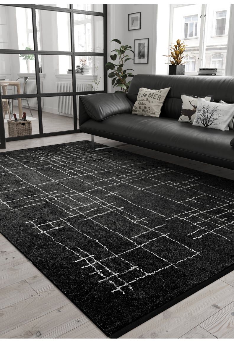 Modern living room with the Bohemian Illusion - Washable Rug - JR1953, black leather sofa with patterned cushions, light wood floor, potted plants, and large windows for natural light. Minimalist decor in monochrome tones.