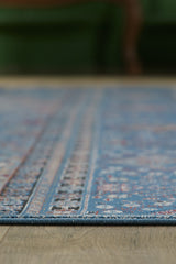 Close-up side view of the Bohemian Tracery Rug - Blue - 2221B, showcasing its blue pattern edge on a wooden floor with a blurred green background.