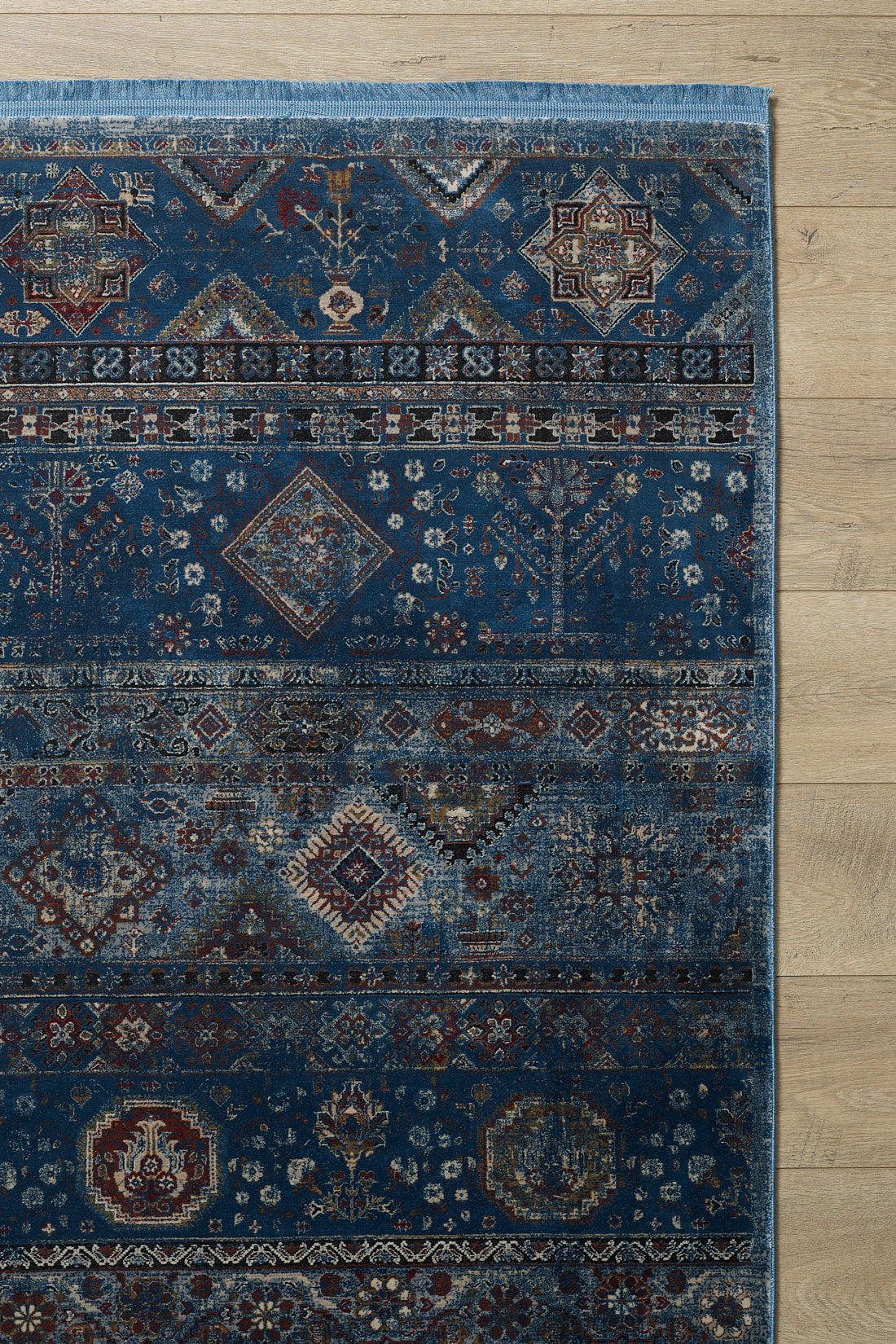 The Bohemian Tracery Rug - Blue - 2221B showcases intricate navy, cream, and burgundy geometric and floral patterns in elegant horizontal bands, making a striking statement on a light wood floor.