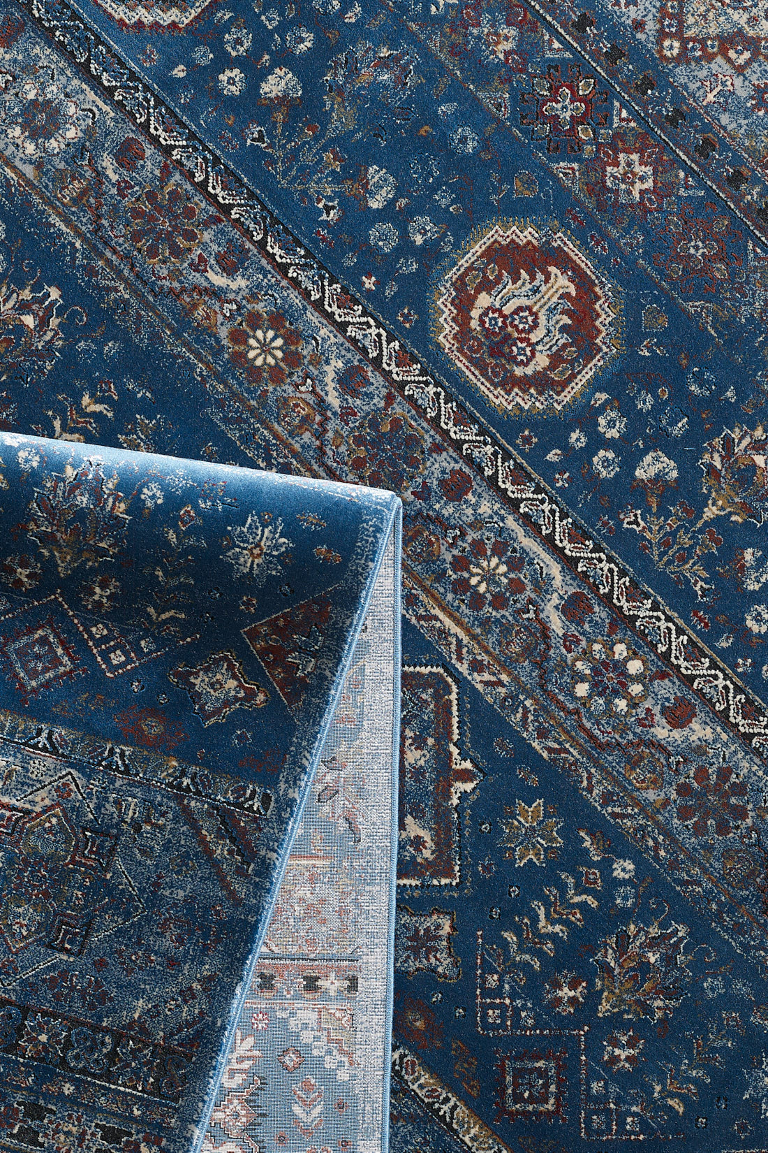 The Bohemian Tracery Rug - Blue - 2221B features intricate floral and geometric patterns in blue, red, white, and gold. One corner is folded over to display the lighter underside and backing.