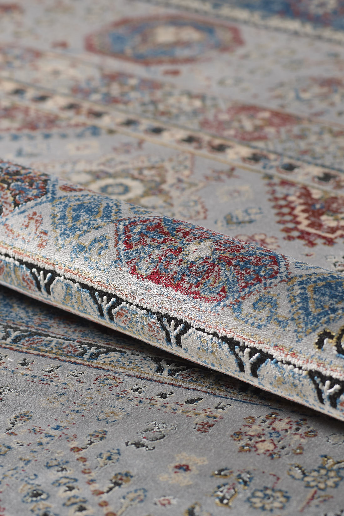 A close-up of the Bohemian Tracery Rug - Grey - 2221C highlights its intricate geometric patterns in blue, red, cream, and beige, showcasing the woven texture and detail with a portion of the rug gently folded.