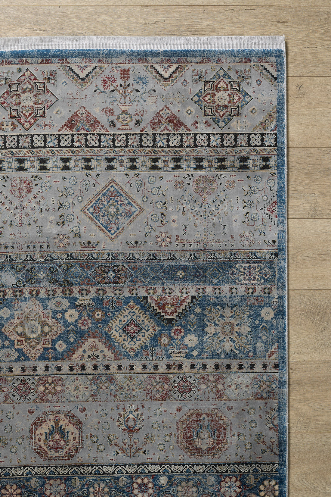 A portion of the Bohemian Tracery Rug - Grey - 2221C, featuring geometric and intricate floral patterns in muted blue and grey tones, is shown on a light wood floor. This elegant machine-made rug adds a traditional touch to the space.