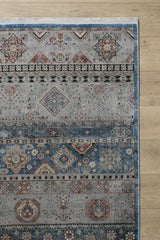 A portion of the Bohemian Tracery Rug - Grey - 2221C, featuring geometric and intricate floral patterns in muted blue and grey tones, is shown on a light wood floor. This elegant machine-made rug adds a traditional touch to the space.