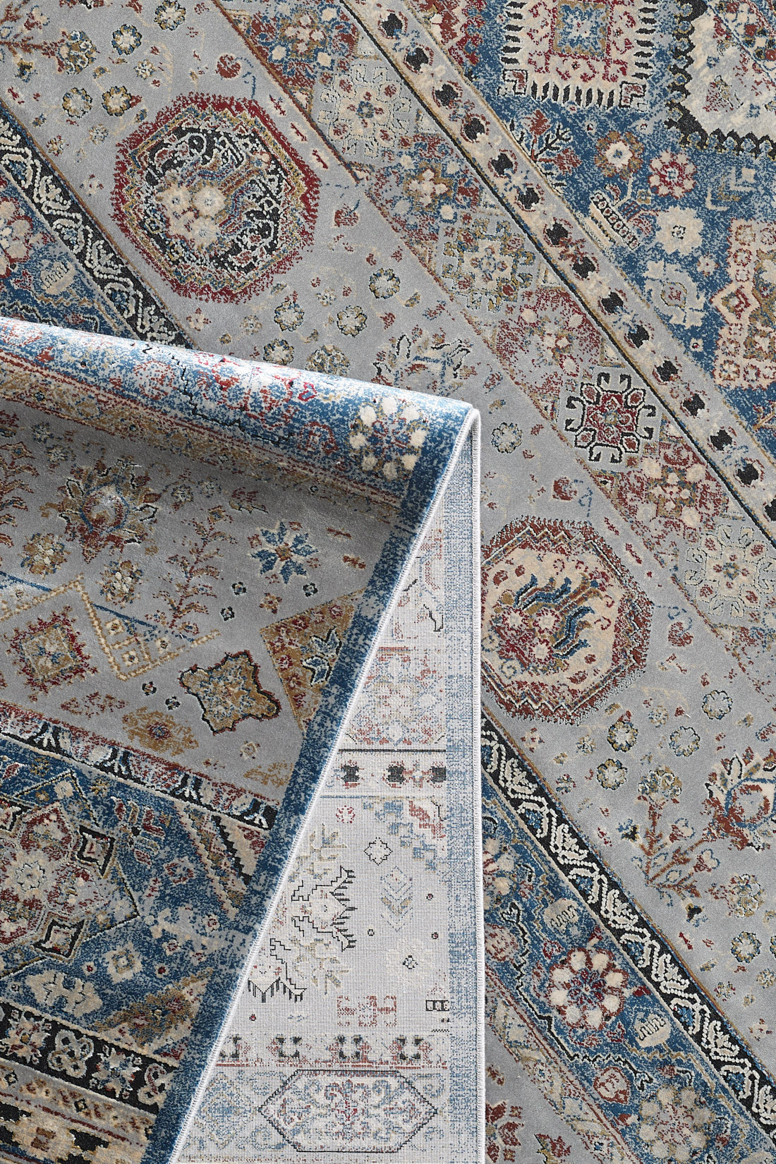 A detailed view of the Bohemian Tracery Rug - Grey - 2221C, a blue and grey machine-made rug with intricate floral and geometric patterns in red, cream, and beige, shown partly folded to display both sides.