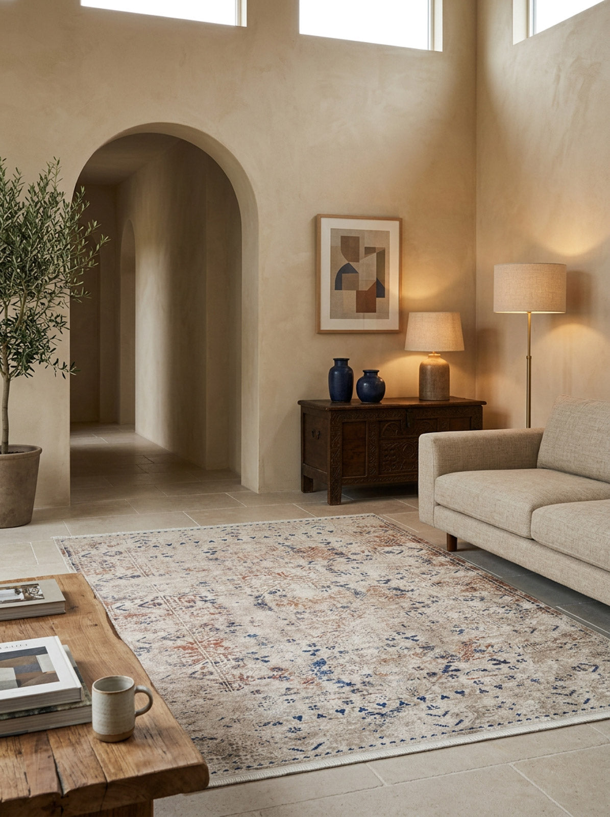 A warm, minimalist living room with beige walls, arched doorway, potted olive tree, neutral sofa, wooden coffee table, console with decor, framed art, two lamps for soft lighting, and a Boho Babe - Washable Rug - JR1664 for high-traffic areas.