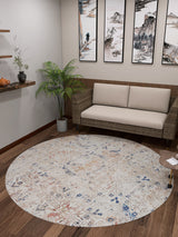 A cozy room featuring the Boho Babe - Washable Rug - JR1664 (Custom Size) in cream and beige on a wooden floor, a wicker sofa with beige cushions, art, shelves with decor, plants, and a small table—ideal for high-traffic areas.