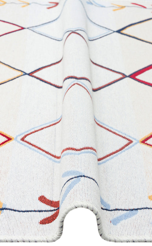Load image 1 in gallery view. A close-up of the Boho Charm - Washable Reversible Kilims - AR21 rug, a white flatwoven piece featuring a colorful geometric pattern in red, blue, yellow, and black, with a raised fold running lengthwise down the center.