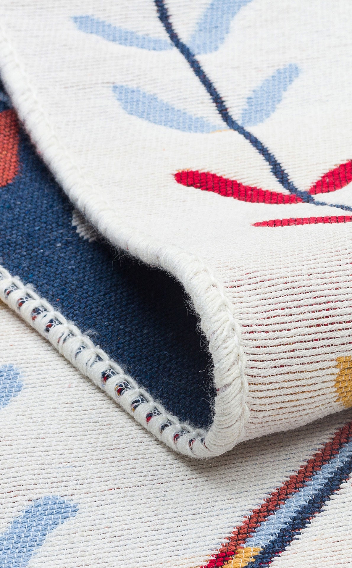 A close-up of the Boho Charm - Washable Reversible Kilims - AR21 rug shows a folded corner with a floral pattern in red, blue, and yellow on a cream background. Visible edge stitching highlights the rug’s intricate threadwork.