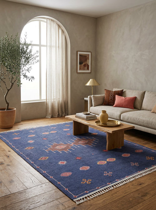 Load image 1 in gallery view. A cozy living room with a beige sofa, colorful cushions, a wooden coffee table, the Boho Chic Tapestry - Washable Rug - JR5083 in blue, a potted plant by an arched window with sheer curtains, and neutral-toned walls.