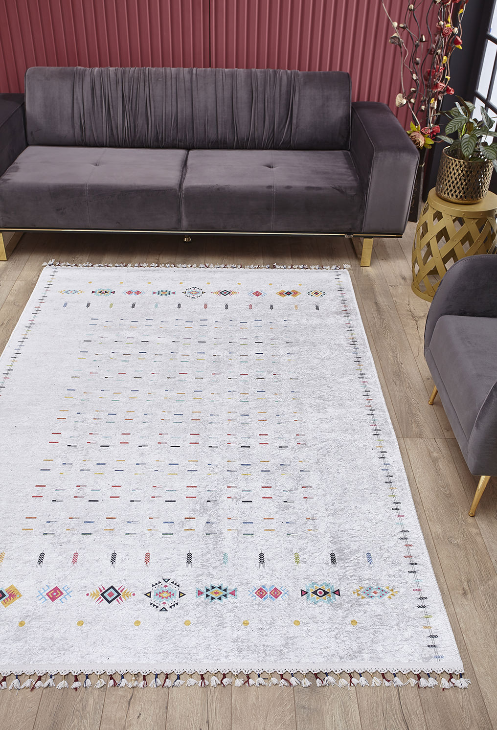 A modern living room with a gray velvet sofa, wood flooring, gold side table, and red vertical panels features the Boho Chic - Washable Rug - JR5103 (Custom Size), adding colorful geometric style and practicality to busy spaces.