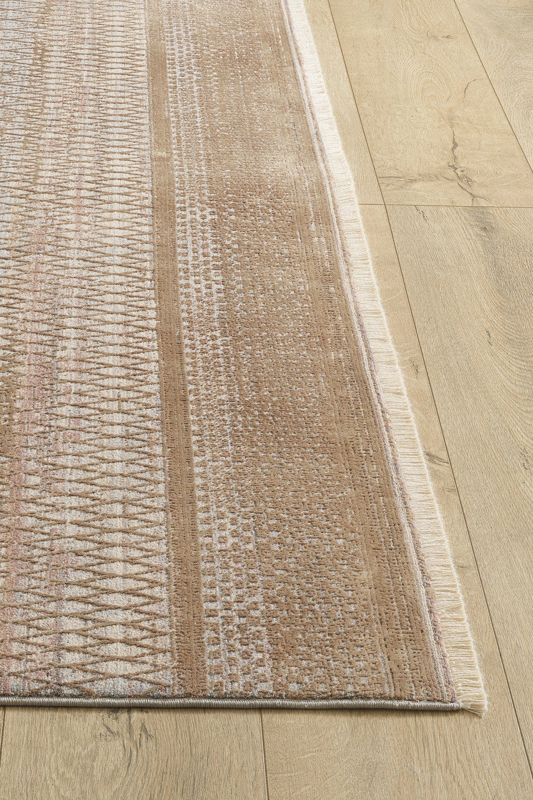 A corner of the Boho Sunset Trellis Rug - HRD004, a machine-made beige and light brown area rug with geometric and linear patterns plus fringed edges, is shown on a light wooden floor.