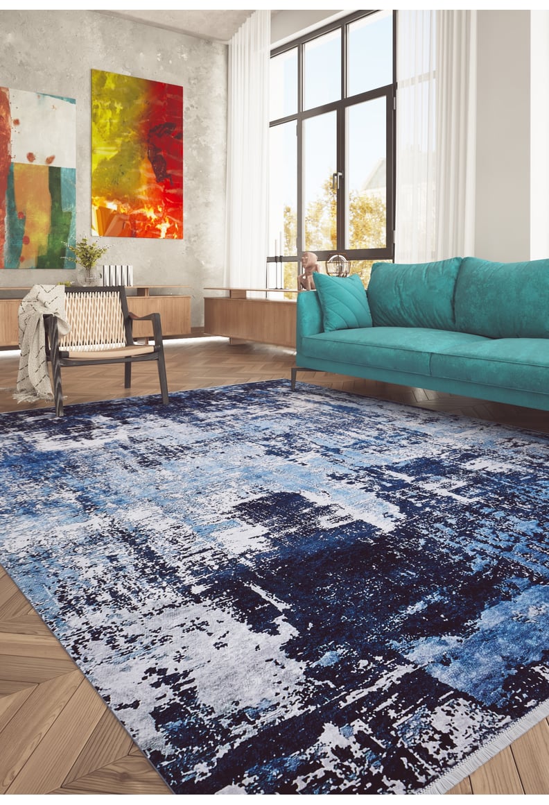 Modern living room featuring the Bold Blooms - Washable Rug - JR836 (Custom Size) in blue, perfect for high-traffic areas, paired with a teal sofa, black chair, wooden flooring, large windows with white curtains, and colorful abstract art on concrete walls.