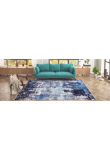 A modern living room features a teal sofa, wooden floors, large windows, and plants on a low shelf. The Bold Blooms - Washable Rug - JR836 (Custom Size) in blue adds style—perfect for high-traffic areas. A beige throw and books rest on a side chair.
