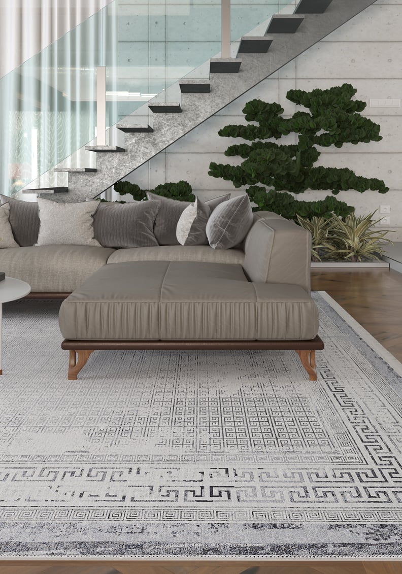 A modern living room with a gray sectional, neutral throw pillows, potted plants, and an open staircase with a glass railing, featuring the Bold Borderline - Washable Rug - JR1984 in gray-anthracite—ideal for high-traffic areas.