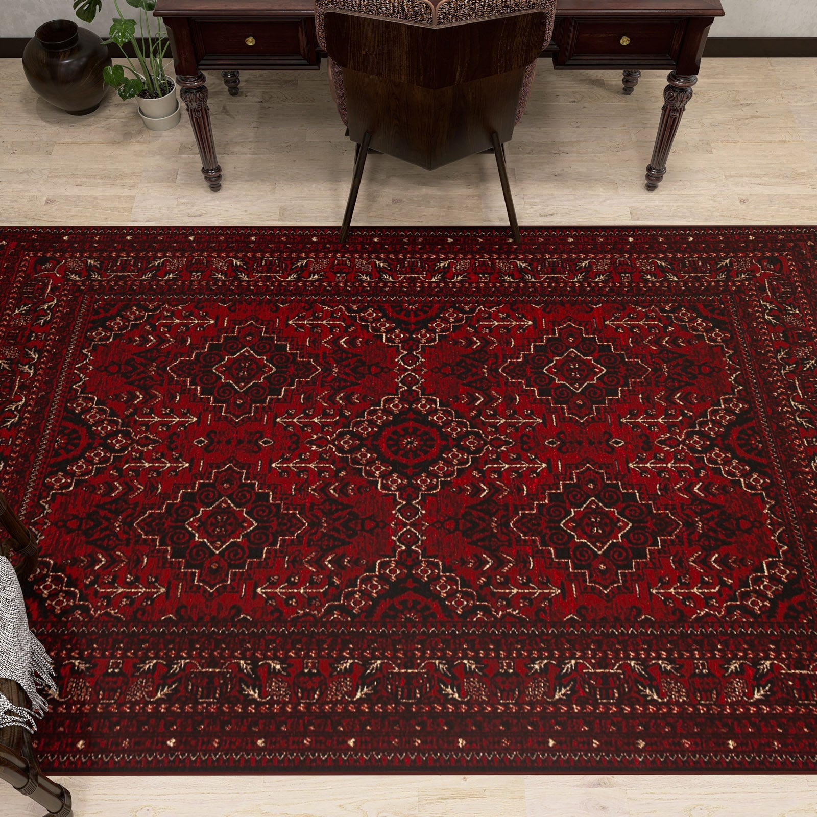 The Bold Thread - Kilim - BHR05 is a rectangular red Turkish rug with geometric and floral patterns, made of soft chenille-polyester fibers. It’s shown on a light wood floor by a dark wooden desk and chair, with a plant nearby.