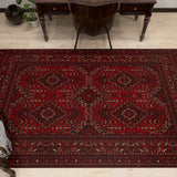 The Bold Thread - Kilim - BHR05 is a rectangular red Turkish rug with geometric and floral patterns, made of soft chenille-polyester fibers. It’s shown on a light wood floor by a dark wooden desk and chair, with a plant nearby.