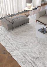 A modern living room features the Borderline Brilliance - Washable Rug - JR1987, ideal for high-traffic areas, paired with a gray chaise lounge, white side table with lamp and book, light gray sofa, and sheer curtains letting in natural light.