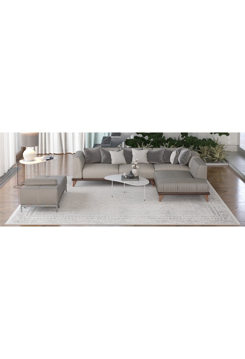 A modern living room features a gray sectional, ottoman, white coffee table, and the Borderline Brilliance - Washable Rug - JR1987 in gray-anthracite, accented by plants near large windows with sheer curtains.