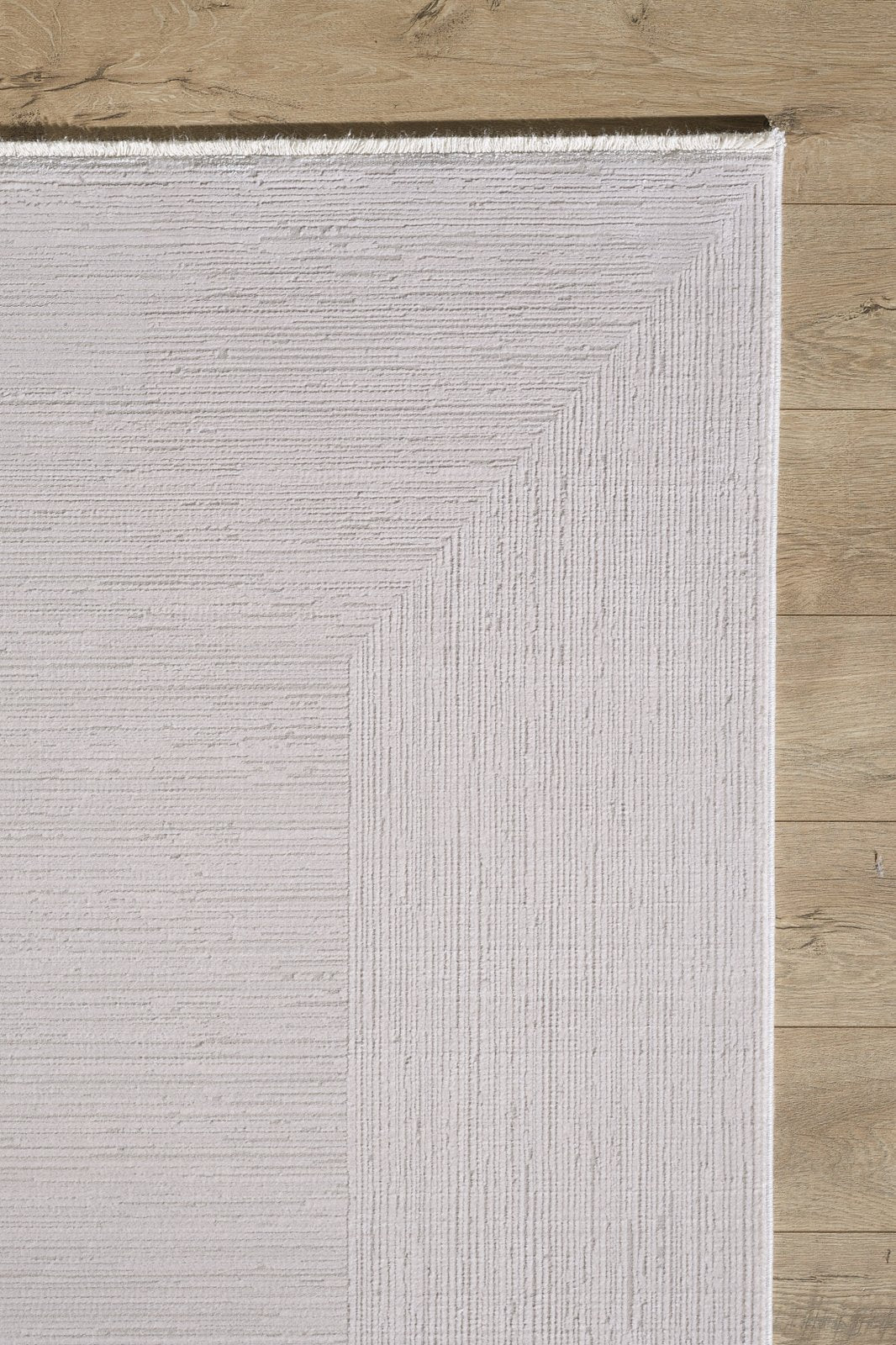 A close-up shows a corner of the Candlelight Comfort Modern Ivory Rug - EW2824 with subtle linear patterns and pale beige tones, placed on a light wood floor. This rectangular rug features a neutral palette and is machine-made.