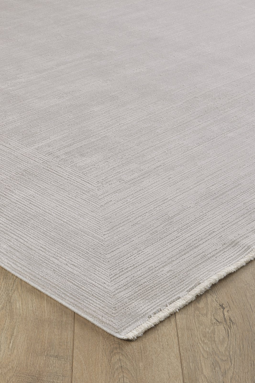 A close-up of the Candlelight Comfort Modern Ivory Rug - EW2824 shows its textured light gray design with subtle horizontal lines and short fringe on one corner, elegantly placed on a wooden floor.