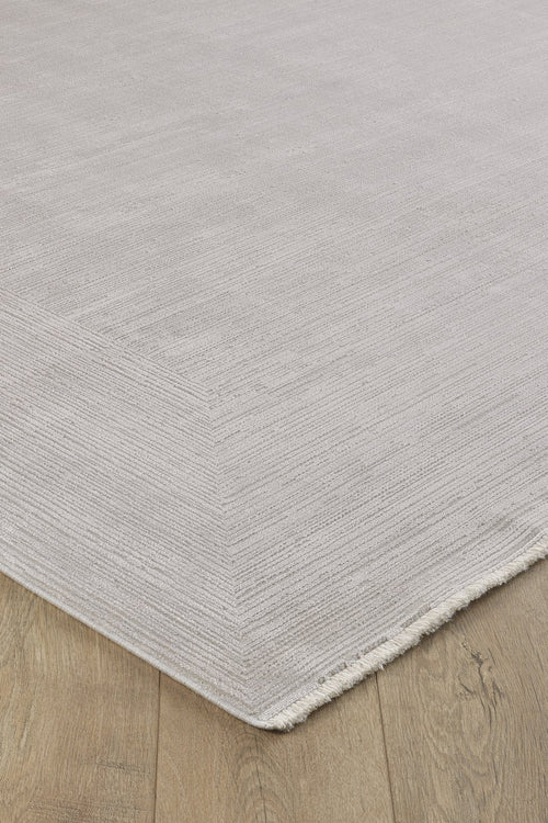 Load image 1 in gallery view. A close-up of the Candlelight Comfort Modern Ivory Rug - EW2824 shows its textured light gray design with subtle horizontal lines and short fringe on one corner, elegantly placed on a wooden floor.
