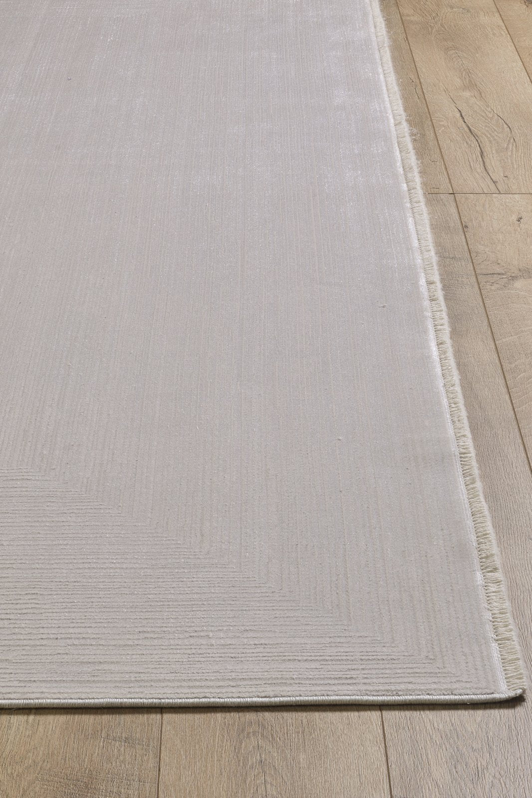 A close-up of the Candlelight Comfort Modern Ivory Rug - EW2824, a machine-made, neutral-toned rug with subtle textured lines and short fringe on one edge, shown atop a warm wooden floor for a soft look.