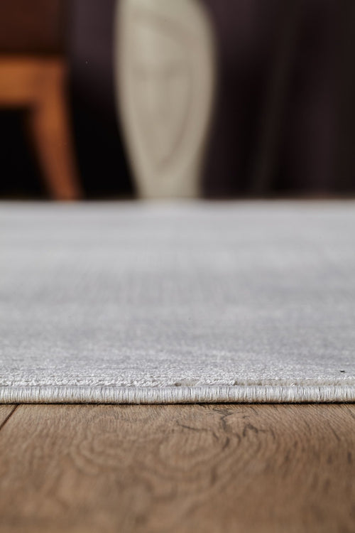 Load image 1 in gallery view. Close-up of the edge of the Candlelight Comfort Modern Ivory Rug - EW2824 on a wooden floor, with blurred furniture and decor in the background.