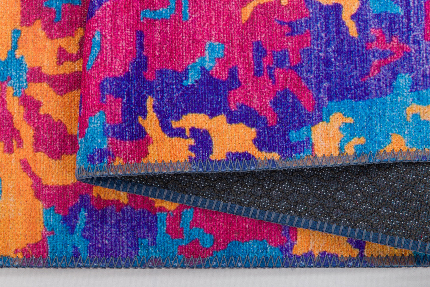 A close-up of the Celestial Tapestry - Washable Rug - JRS004 (Custom Size), featuring a colorful abstract pattern in pink, orange, blue, and purple. The folded edge reveals a dark underside with blue stitching—great for allergy-sensitive or busy spaces.