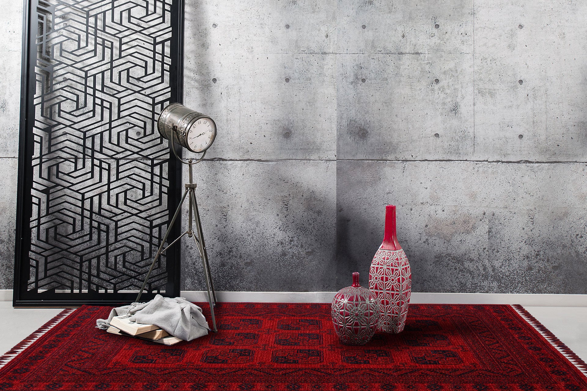The Cherry Texture - Kilim - BHR01, in cherry red with Turkish patterns, is placed on a light floor, accompanied by three vases, a tripod spotlight, folded fabric, books, and a geometric black screen against a concrete wall.