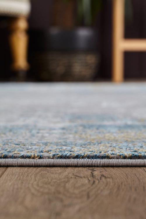 Load image 1 in gallery view. A close-up of the ChromaBurst Premium Rug - Blue - MT176, showing its modern blue and gray edge on a wooden floor, with blurred furniture and a planter in the background.