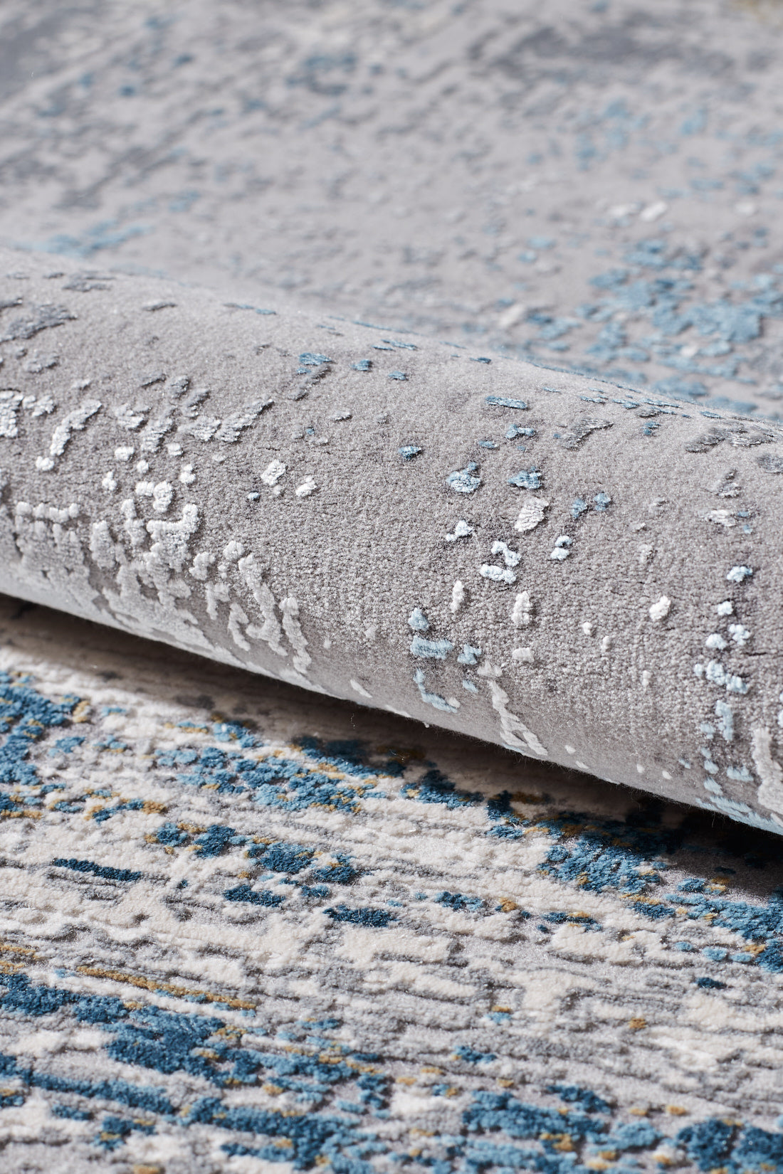 Close-up of the ChromaBurst Premium Rug - Blue - MT176, partially rolled to reveal its soft texture and abstract gray and blue design—this machine-made rug features a stylish pattern in blue, gray, and off-white.