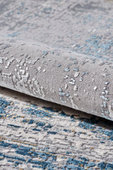 Close-up of the ChromaBurst Premium Rug - Blue - MT176, partially rolled to reveal its soft texture and abstract gray and blue design—this machine-made rug features a stylish pattern in blue, gray, and off-white.