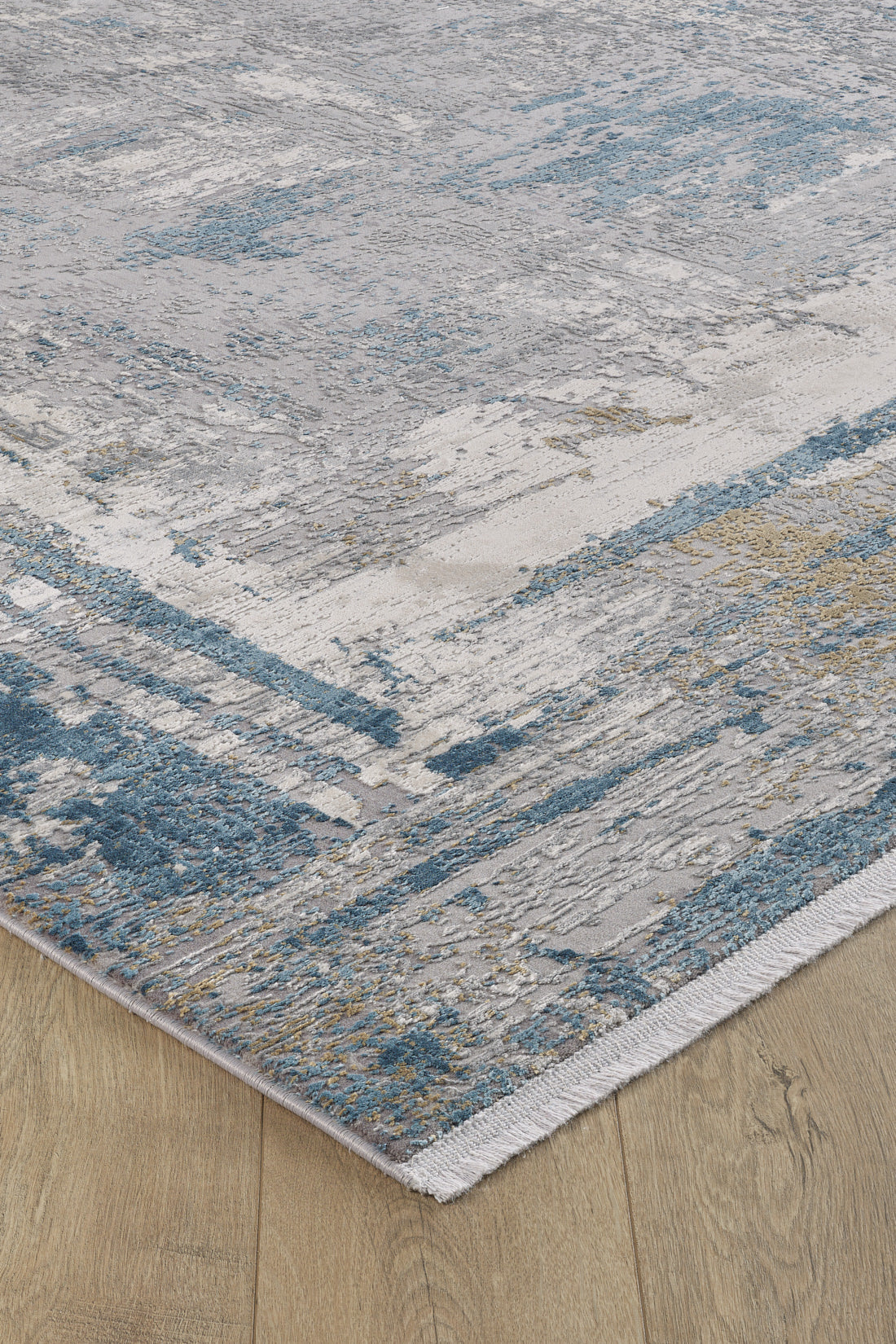A close-up of the ChromaBurst Premium Rug - Blue - MT176, a machine-made rug with a distressed abstract pattern in blue, gray, beige, and white on a light wood floor. It features a visible woven edge and textured appearance.