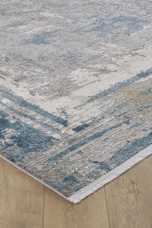 Load image 1 in gallery view. A close-up of the ChromaBurst Premium Rug - Blue - MT176, a machine-made rug with a distressed abstract pattern in blue, gray, beige, and white on a light wood floor. It features a visible woven edge and textured appearance.
