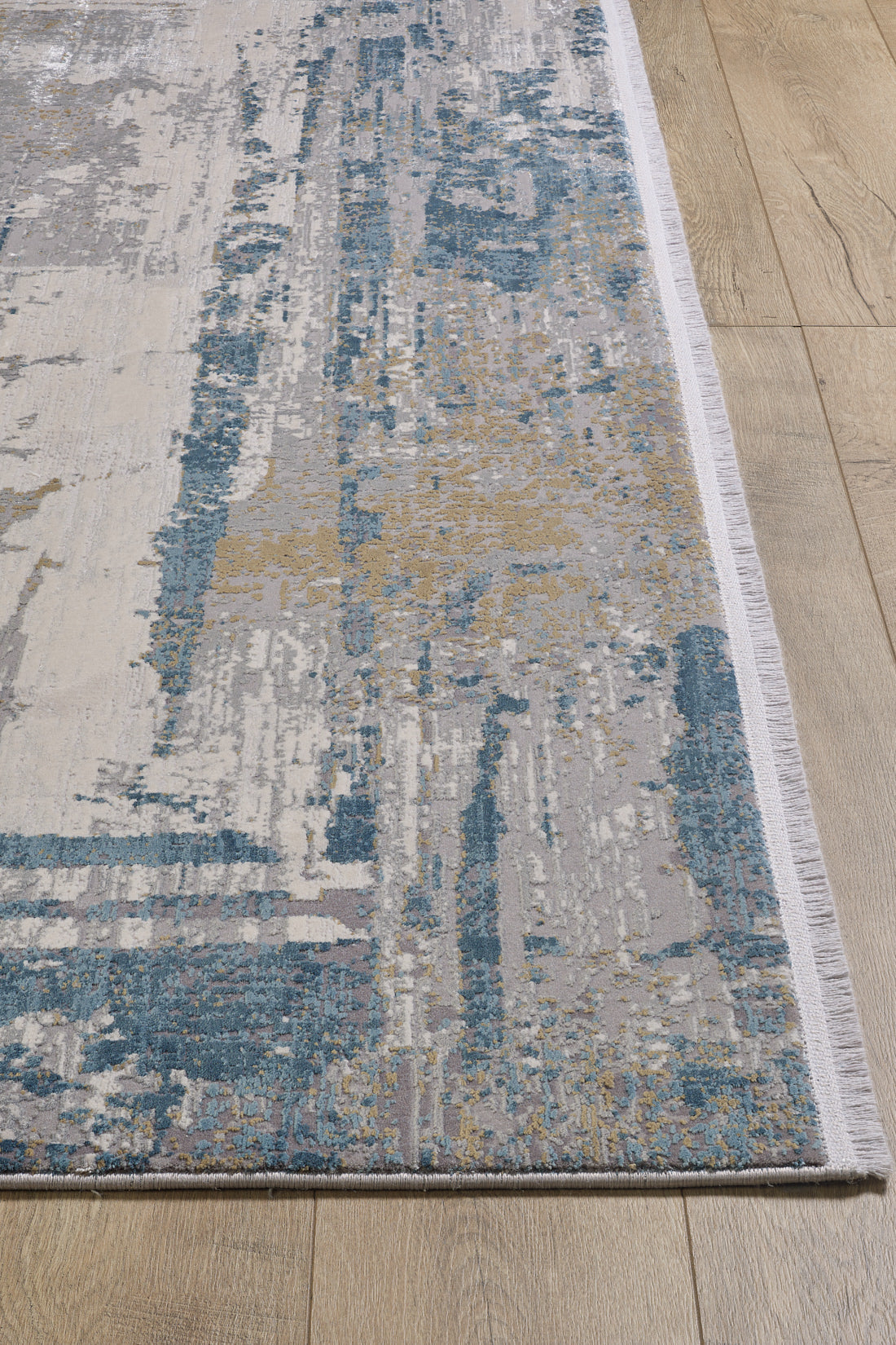A close-up of the ChromaBurst Premium Rug - Blue - MT176, a modern machine-made acrylic-viscose rug with an abstract blue, beige, gray, and white pattern and a white fringe, displayed on a light wooden floor.