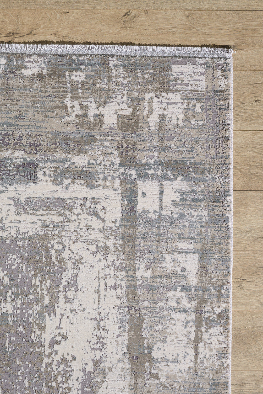 A close-up of the ChromaBurst Premium Rug - Bright - MT177, a modern machine-made rug in light gray and cream with an abstract distressed pattern and visible fringe, placed on a light wood floor.