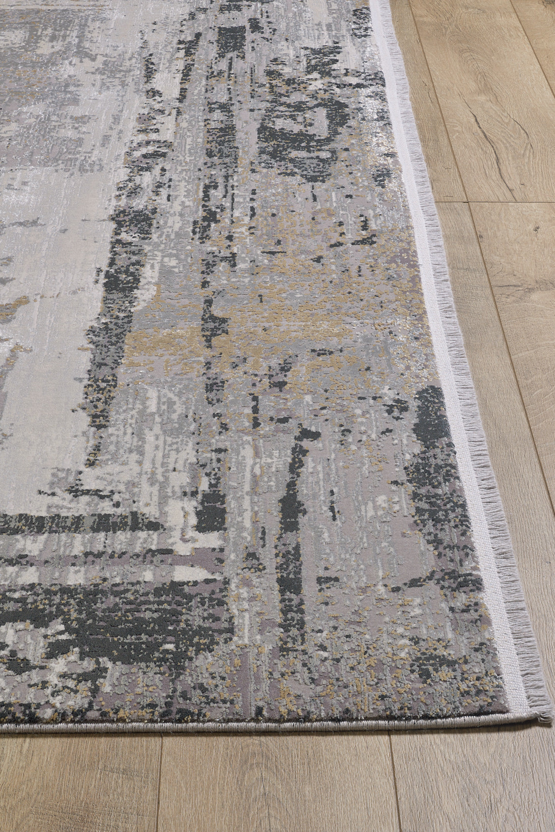 A close-up of the ChromaBurst Premium Rug - Dark - MT175 shows its abstract gray, beige, and gold pattern on a light wood floor. The machine-made rug features a white fringe along the edge for a stylish finish.