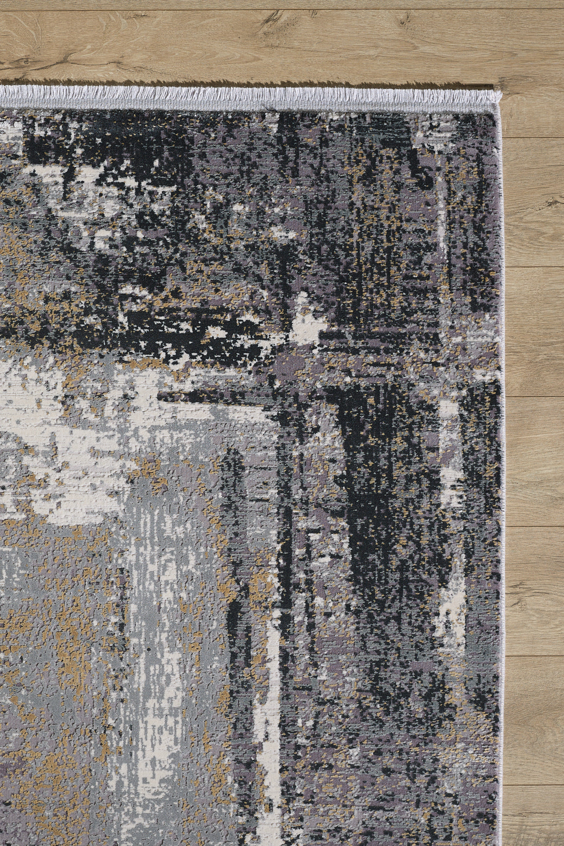 A close-up of the ChromaBurst Premium Rug - Dark - MT175 shows its modern abstract pattern in gray, black, beige, and white shades with fringe along the upper edge, resting on a light wooden floor.