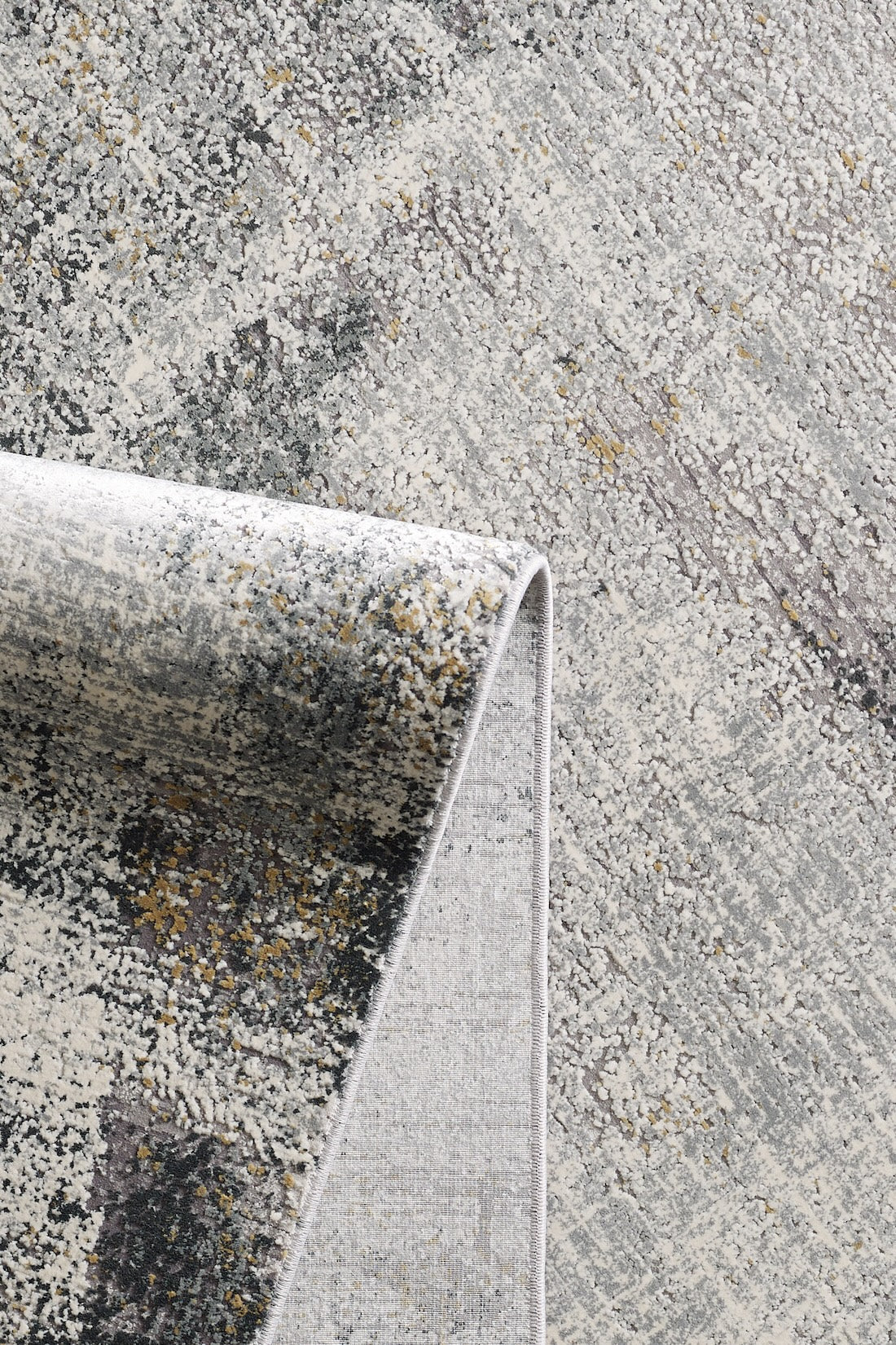 A close-up of the Chromatic Flair Contemporary Rug - Charcoal (M660A) shows its abstract, textured pattern in gray, cream, and gold hues with a soft woven look and a folded corner.