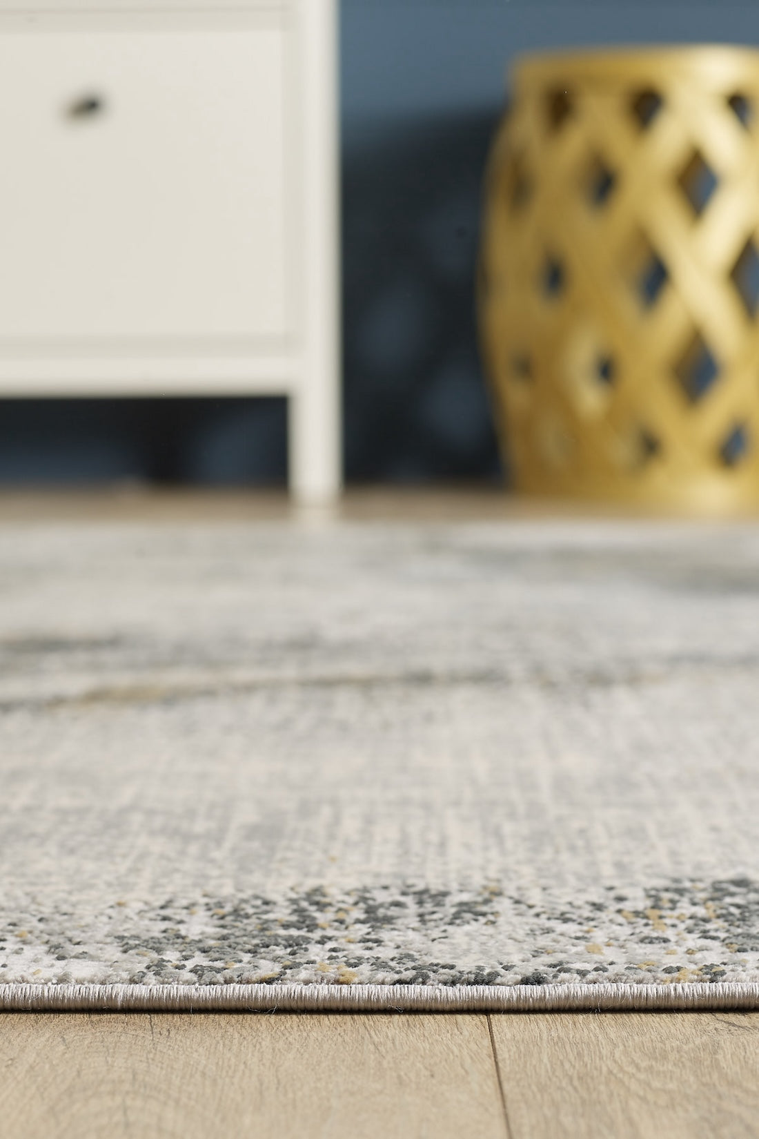 A close-up of the Chromatic Flair Contemporary Rug - Charcoal - M660A on a wooden floor, with a white cabinet and a decorative gold stool softly blurred in the background.