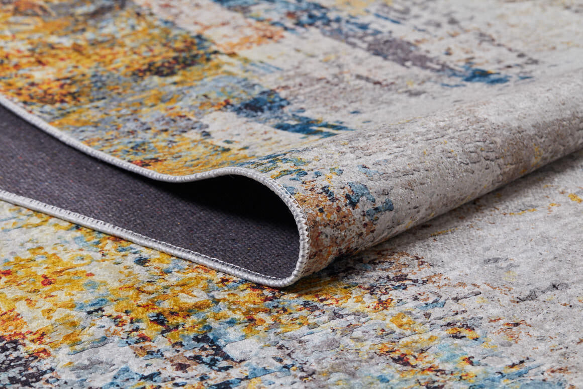 A close-up of the Chromatic Kaleidoscope - Washable Rug - JR1483 (Custom Size) in yellow-gold with an abstract blue, orange, and gray pattern. One corner is folded to reveal the dark backing—perfect for high-traffic areas.