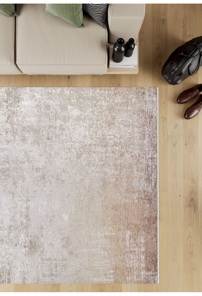 A top-down view of a beige sofa with green pillows sits beside the Cinnamon Swirl - Washable Rug - JR425 (Custom Size) on a wooden floor, near a black bag, brown shoes, and two bottles atop a small stack of books.