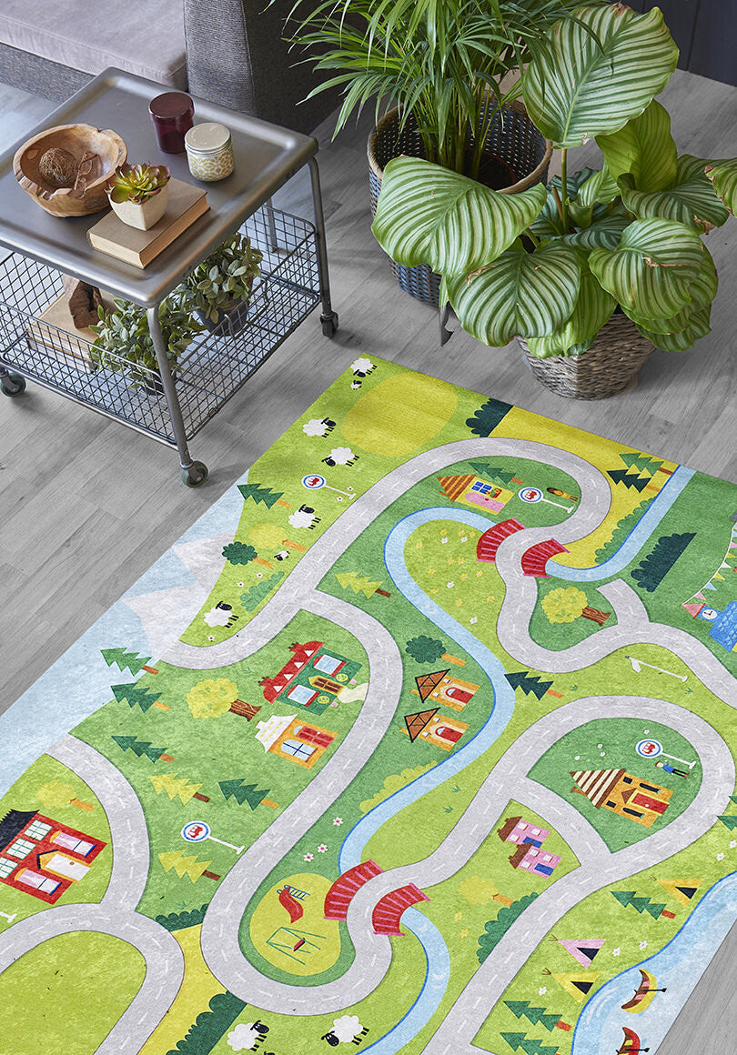The City Adventure - Machine-Washable Rug JRC039 (Custom Size) adds color with winding roads, rivers, houses, trees, and animals—ideal for high-traffic areas—and is shown on a wooden floor beside a metal table with snacks and plants.