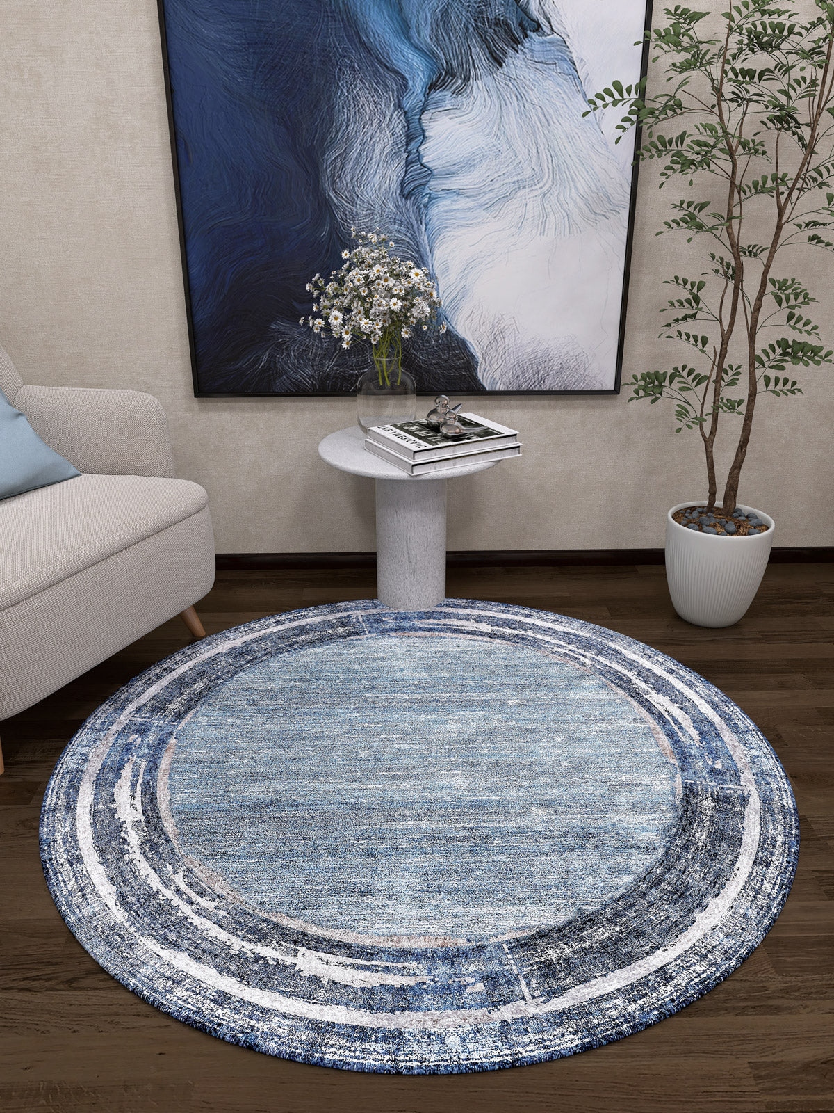 A modern living room corner with the Classic Bordered - Washable Rug - JR1197 (Custom Size) in blue, perfect for high-traffic areas, a small white side table with white flowers, a leafy potted plant, a beige sofa, and abstract blue wall art.
